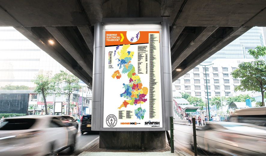 Colourful map of the United Kingdom on a billboard in the middle of a busy urban street, under a concrete bridge. There are cars going past on the left and people can be seen in the distance on the right