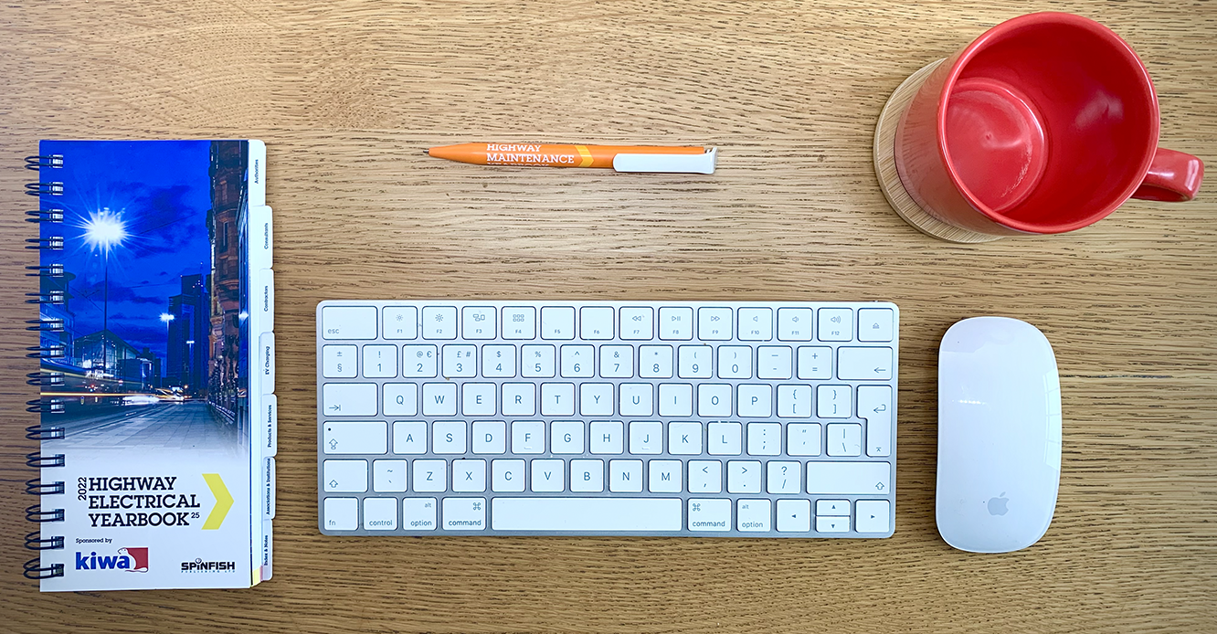A keyboard on a wooden desk with a Highway Electrical Yearbook to the left of it. On the right is a mouse and a red mug and above the keyboard is an orange pen