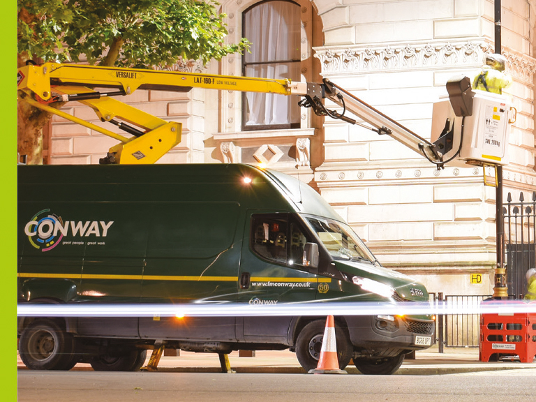 Versalift van mounted access platform attached to FM Conway van. Person in raised bucket working on lamppost, while another person works on the ground surrounded by protective barriers. Traffic cones surround the vehicle for safety