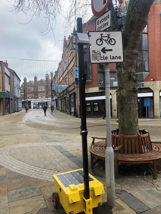 Quiet town centre street with a bench around a tree trunk. Air quality monitor attached to a black post. Cycle lane signage on a post beside it